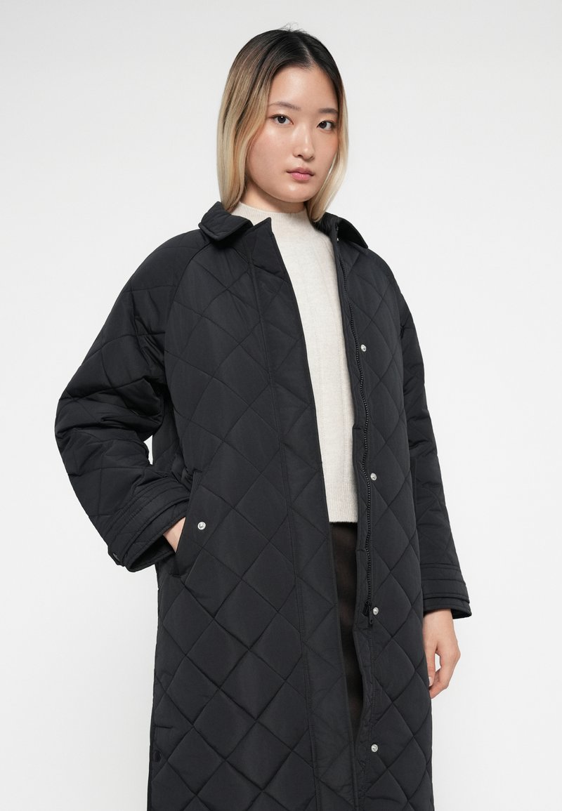 Gequilte zwarte lange jas met een kraag, drukknoopsluitingen en zijzakken. De stof heeft een gladde textuur met ruitpatronen.