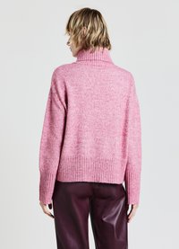 Rosa gestrickter Pullover mit hohem, geripptem Kragen und geripptem Saum, der ein strukturiertes Muster und abgesenkte Schultern aufweist. Trägt dazu eine dunkle Hose.