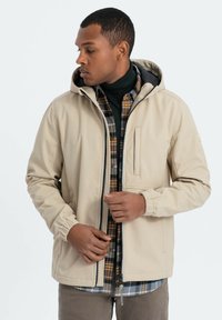 Beige vattentät jacka med huva, elastiska ärmmuddar och dragkedja fram. Bärs över en rutiga skjorta i gröna, gula och blå toner.