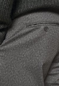 Tissu texturé gris de pantalons présentant une poche avec un bouton, exposé aux côtés d'un vêtement en maille gris foncé.
