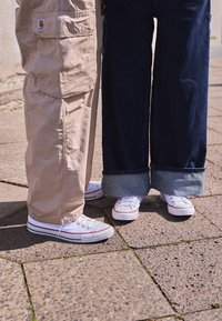 Beżowe spodnie cargo i granatowe dżinsy o szerokich nogawkach, oba zestawione z białymi tenisówkami z czerwonymi akcentami i teksturowanymi podeszwami, na kamiennej nawierzchni.