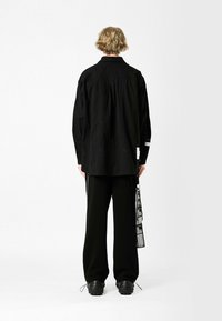 Camisa negra con patrones bordados sutiles, de mangas largas y ajuste holgado. Combinada con pantalones anchos negros y zapatos negros gruesos.