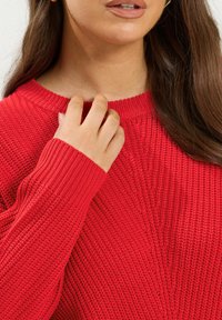 Pull en tricot rouge avec un col rond côtelé et des manches longues. La texture est épaisse avec un motif subtil le long des épaules.