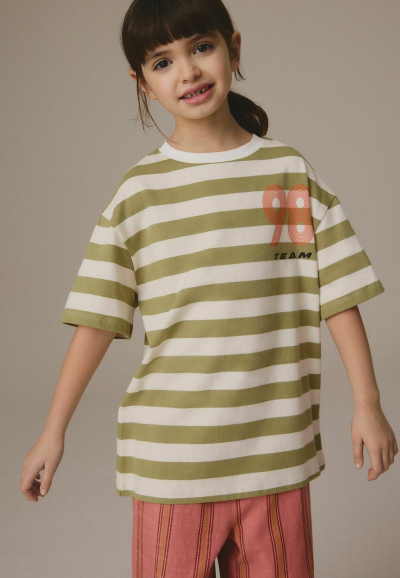 Kind mit dunklen Haaren, das ein grün-weiß gestreiftes T-Shirt mit dem Text "98 TEAM" und rosa gestreifte Hosen trägt, steht vor einem einfarbigen Hintergrund.