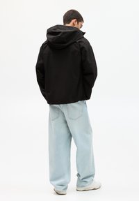 Schwarze Kapuzenjacke mit lockerer Passform, kombiniert mit hellblauen weit geschnittenen Jeans und weißen Turnschuhen, von hinten betrachtet.