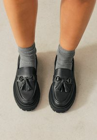 Mocasines de cuero negro con un acabado texturizado, que presentan un detalle decorativo de borlas y hebilla. Combinados con calcetines tobilleros grises.