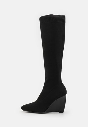 Bottes noires jusqu'au genou avec un dessus en tissu texturé, un bout pointu et un talon compensé. Couleur unie avec un détail de poignet côtelé.