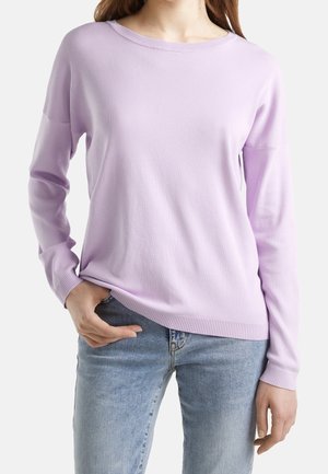 Kvinde iført en lyse lilla langærmet sweater og lyseblå jeans, med den ene hånd delvist i en forlomme.