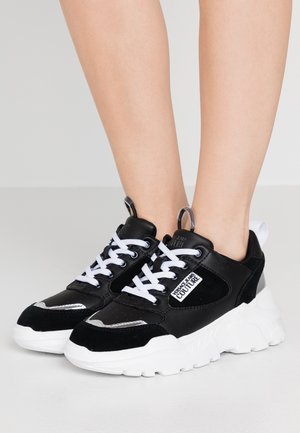 Feet wearing black and white chunky sneakers with white laces and "Versace Jeans Couture" logo on the side.