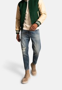 Green and beige varsity jacket with striped cuffs, white sweatshirt, distressed light blue jeans, and tan lace-up boots.