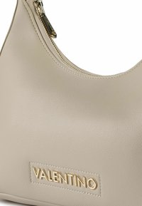 Borsa a tracolla in pelle beige con forma arrotondata, dettaglio con cerniera dorata e finitura strutturata. Presenta il logo "Valentino" in rilievo sulla parte anteriore.