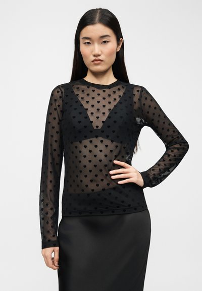 Νεαρή γυναίκα με μακριά σκούρα μαλλιά, φορώντας διάφανο μαύρο μακρυμάνικο μπλουζάκι με μικρά σχέδια καρδιών και μαύρο bralette από κάτω.