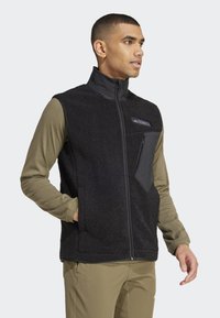 Schwarze Fleece-Weste mit hohem Kragen, Reißverschluss vorne und einer Brusttasche. Getragen über einem langärmligen olivgrünen Shirt mit eng anliegenden Ärmeln.