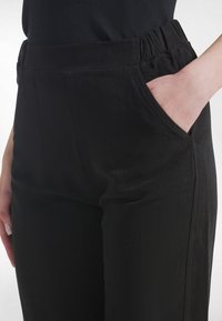 Mustat joustovyötäröiset housut, joissa on sivutaskut, sileä pinta, suora lahjemalli ja sivusaumoissa yksityiskohtia.