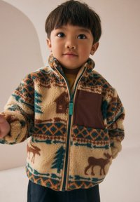 Jeune enfant avec des cheveux courts et foncés portant une veste en polaire à motifs avec des designs d'arbres et d'élans, se tenant devant un fond neutre.