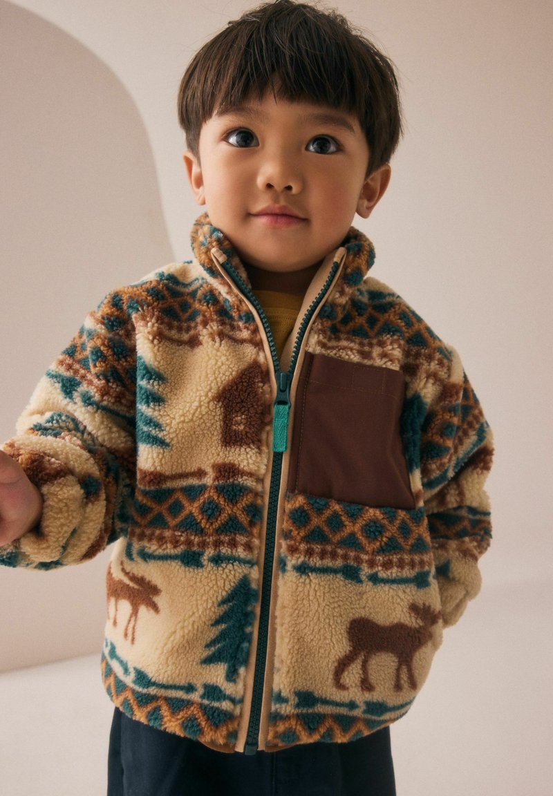 Jeune enfant avec des cheveux courts et foncés portant une veste en polaire à motifs avec des designs d'arbres et d'élans, se tenant devant un fond neutre.