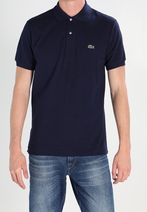 Mann trägt ein dunkelblaues Lacoste-Poloshirt mit zwei Knöpfen und hellblauen Jeans, steht mit entspannten Armen an den Seiten.
