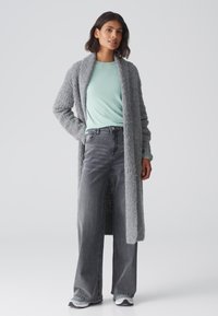 Grijze, gestructureerde lange cardigan over een lichtblauw shirt, gecombineerd met hoge grijze jeans en grijze sneakers. Eenvoudig, casual ontwerp.