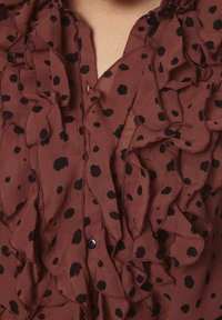 Brązowa przezroczysta bluzka z czarnymi kropkami, z falbanami przy dekolcie i listwie guzikowej, z widocznymi detalami szycia.