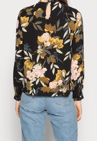 Blouse noire à motifs floraux avec manches longues, ornée de fleurs roses et jaunes, de feuilles vertes, et une découpe en forme de trou de serrure au dos, portée avec un jean denim bleu.