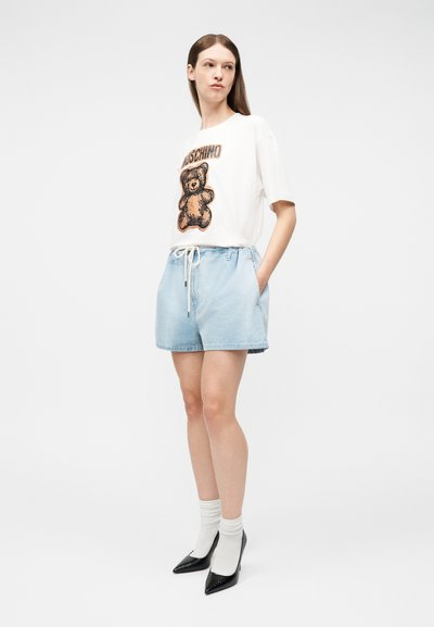 Γυναίκα με λευκό t-shirt με σχέδιο αρκουδάκι, ανοιχτομπλε σορτς με κορδόνι, λευκές κάλτσες και μαύρα μυτερά τακούνια, στέκεται με τα χέρια στις τσέπες.