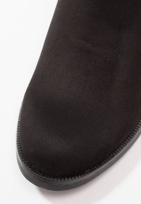 Gros plan avant d'une chaussure en daim noir avec un bout arrondi et une semelle noire cousue sur fond blanc.
