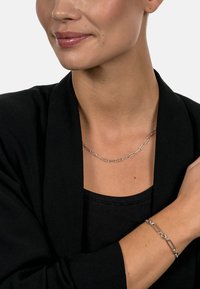 Silver chain necklace with irregular link shapes and bracelet with similar design, both worn over a black top and blazer.