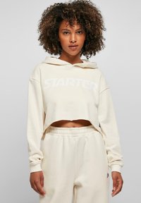 Sweat à capuche crème court avec un logo "STARTER" blanc, doté de manches longues et d'une capuche avec cordon ; associé à un pantalon de survêtement crème assorti.