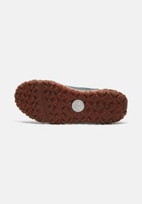 Suela de goma con treads multidireccionales en marrón, que presenta un logo circular plateado. La parte superior muestra una superficie oscura y texturizada.