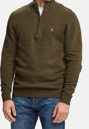 Mann trägt einen olivgrünen Strickpullover mit Viertel-Reißverschluss und kleinem roten Logo auf der linken Brust, kombiniert mit blauen Jeans.
