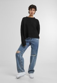 Zwarte geribbelde gebreide trui met een ontspannen pasvorm, gecombineerd met lichtblauwe versleten wijde jeans en witte sneakers. Eenvoudige en casual uitstraling.