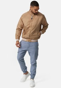 Chaqueta bomber beige con puños y cuello de rayas blancas, combinada con pantalones cargos ligeros en azul claro y zapatillas blancas. Textura de tela suave.