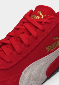 Κοντινή λήψη ενός κόκκινου sneaker Puma από σουέντ με κόκκινα κορδόνια, μια λευκή πλαϊνή ρίγα και χρυσό λογότυπο Puma στη γλώσσα και στο πλάι.