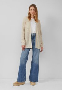 Beige gestrickte Strickjacke über einem weißen T-Shirt, kombiniert mit hoch taillierten blauen Schlagjeans und hellbeigen Plateaustiefeln.