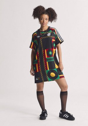 Mladá žena s kučeravými vlasmi v farebných šatách s potlačou a čiernych teniskách stojaca na jednoliatom pozadí s jednou nohou mierne pokrčenou.