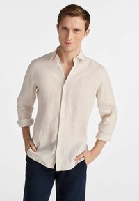 Jasno beżowa lniana koszula z kołnierzykiem zapinanym na guziki i podwiniętymi rękawami, z delikatną fakturą i małym logo na piersi.
