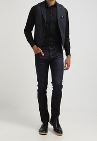Svart skjorta med knappar, grå väst med ficka, mörka jeans och svarta skor med kontrasterande sula. Slim fit, mjuk textur, skräddarsydd design.