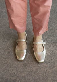 Metalizuoti auksiniai Mary Jane batai su kvadratiniu pirštu ir kulkšnies juosta, derinami su šviesiai rožinėmis trumpomis kelnėmis, parodantys sklandžią tekstūrą ir blizgesį.