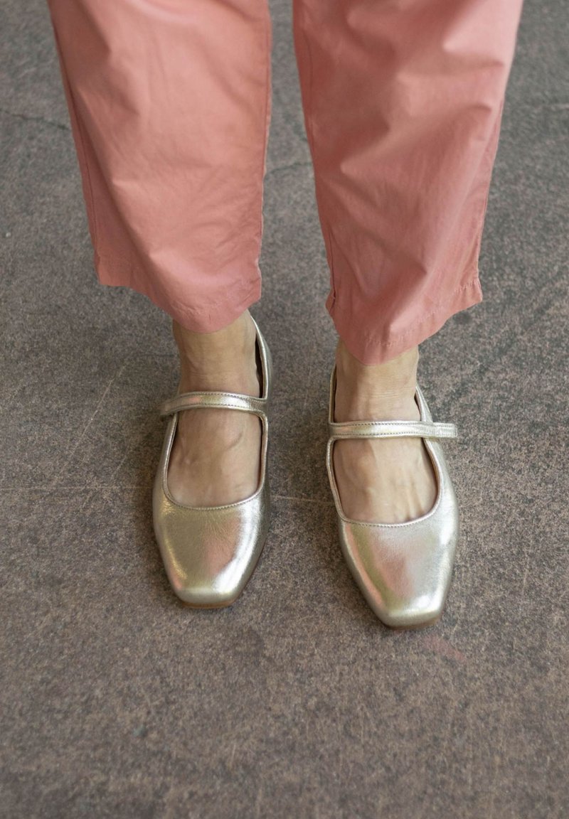 Metalizuoti auksiniai Mary Jane batai su kvadratiniu pirštu ir kulkšnies juosta, derinami su šviesiai rožinėmis trumpomis kelnėmis, parodantys sklandžią tekstūrą ir blizgesį.
