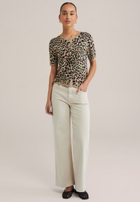 Cardigan à manches courtes à imprimé léopard dans les tons beige et noir, associé à un jean large couleur écru. Le modèle présente une coupe décontractée et des poches avant.