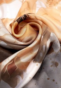 Zijdeachtige stof gevouwen in een spiraalpatroon met abstracte vormen en zachte tinten van beige, bruin en grijs, toont een gladde textuur en lichtreflectie.