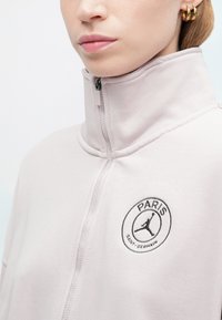 Felpa zip-up rosa chiaro con colletto alto, caratterizzata da un logo rotondo "PARIS" con una silhouette sul lato sinistro del petto. Texture morbida.