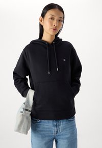 Felpa nera con cappuccio e tasca a marsupio frontale, con aglet argentati sui lacci. Indossata con jeans blu chiaro e una borsa morbida.