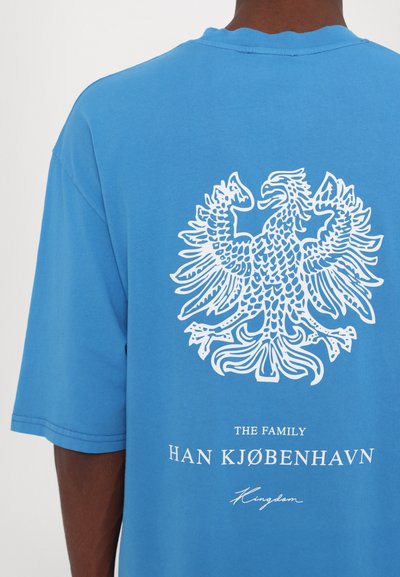 Blå kortärmad t-shirt med en vit design av en dubbelhövdad örn och texten "THE FAMILY HAN KJØBENHAVN Kingdom" under.
