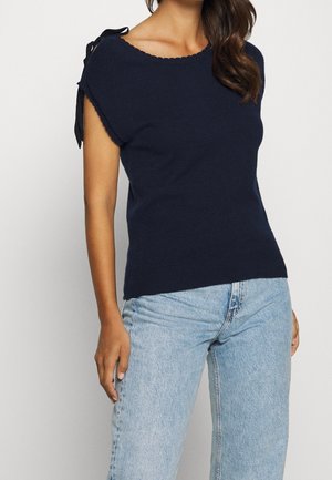 Granatowy sweter z krótkim rękawem i karbowanym dekoltem; ozdobiony czarnymi wiązaniami na ramionach; zestawiony z jasnoniebieskimi dżinsami.