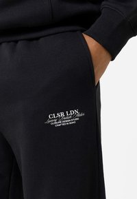 Χέρι ατόμου στην τσέπη μαύρου παντελονιού φόρμας με λευκό λογότυπο κειμένου που αναγράφει "CLSR LDN Luxury British Atelier Closure Design Studio Crafted in Soho."