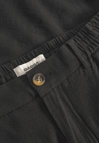 Black woven pants featuring an elastic waistband, a single brown button, and a label reading "GABBA." Smooth, textured fabric detail visible.