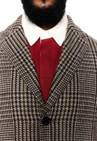 Bārda vīrietis, kas valkā brūnu svītrainu žaketi virs sarkanas krekla ar baltu pogātu apkakli uz baltas fona.