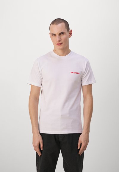 Camiseta blanca de algodón de mangas cortas, cuello redondo y texto bordado en rojo "THE KOOPLES" en el lado izquierdo. Llevada con jeans oscuros.