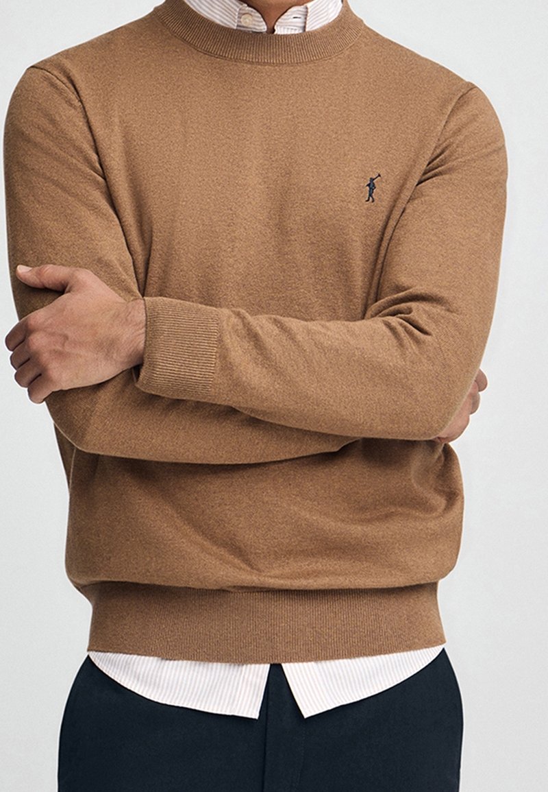 Homme bras croisés portant un pull de couleur chameau sur une chemise blanche boutonnée et un pantalon foncé, montrant un petit logo brodé.
